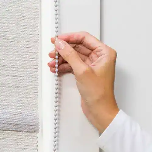 Closely view of a hand pulling down a white bead chain to operate a window blind or shade.