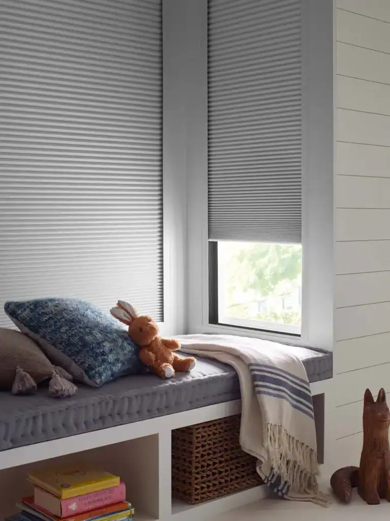 Soft gray window blinds with a cozy window seat adorned with plush pillows and a stuffed bunny, creating a relaxing nook in a bright, modern space perfect for reading or lounging.
