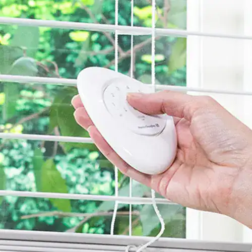 Wireless remote control operating window blinds with a lush green garden background.