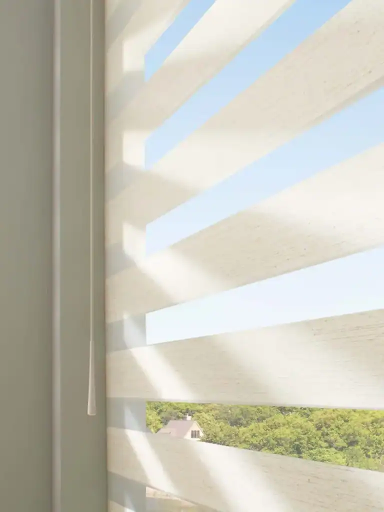 Sunlight filtering through beige vertical blinds with a view of a green treeline and house outside, highlighting the energy-efficient and stylish window covering solution by Superior Blinds.