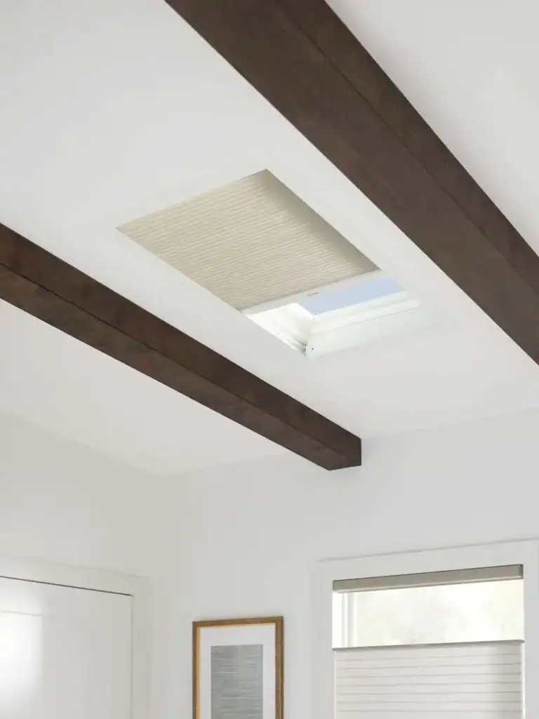 Ceiling skylight with a built-in beige pleated window blind, surrounded by dark wood beams, natural light, and modern interior decor.