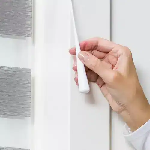 Deluxe white window blinds being installed on a white window frame with a hand holding a white tool, showcasing premium window treatment solutions for home or office.